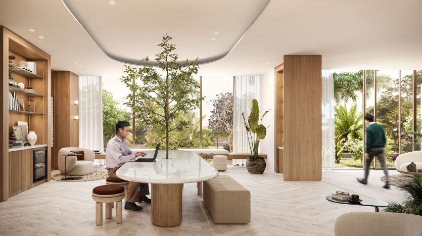 Un salon de bureau moderne dans une agence immobilière de Dubaï avec de grandes fenêtres, des plantes d'intérieur et de la lumière naturelle. Un homme travaille sur un ordinateur portable devant une table en marbre, un autre marche en arrière-plan.