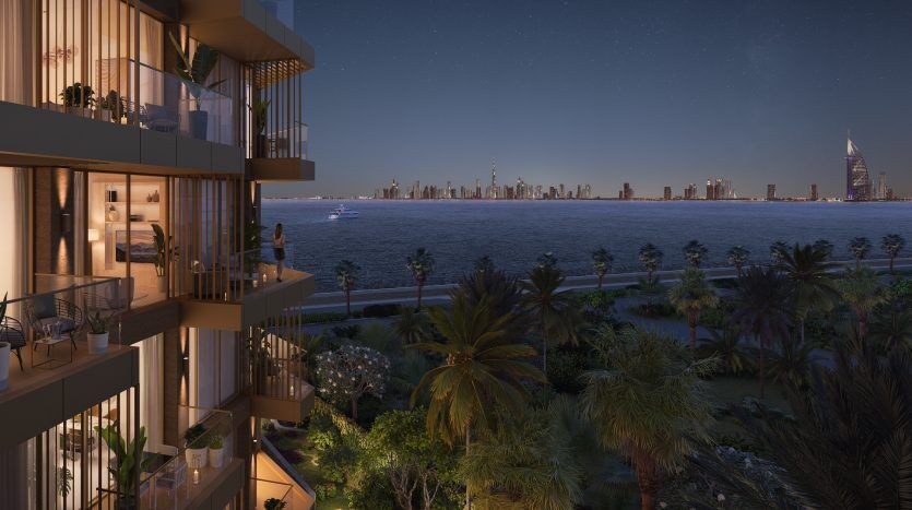 Vue nocturne d'un immeuble d'appartements haut de gamme à Dubaï surplombant les toits de la ville et une mer calme, avec des balcons éclairés chaleureusement et des palmiers au premier plan.