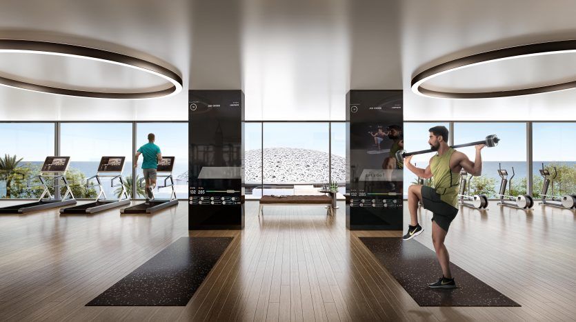 Une salle de sport moderne avec de grandes fenêtres et une vue sur l'océan à Dubaï. Plusieurs appareils d'exercice et deux hommes s'entraînant ; l’un courant sur un tapis roulant et l’autre soulevant des poids.