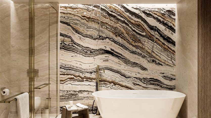 Une salle de bains luxueuse dans une villa à Dubaï, dotée d'une baignoire blanche sur pied, d'une grande douche en verre et de murs recouverts de motifs de marbre ondulés multicolores. Un éclairage doux rehausse l’ambiance élégante.