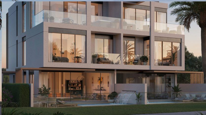 Immeuble résidentiel moderne de plusieurs étages au crépuscule à Dubaï, doté de grandes fenêtres en verre et de balcons avec un mobilier intérieur visible et un éclairage chaleureux. Jardin paysagé au premier plan.