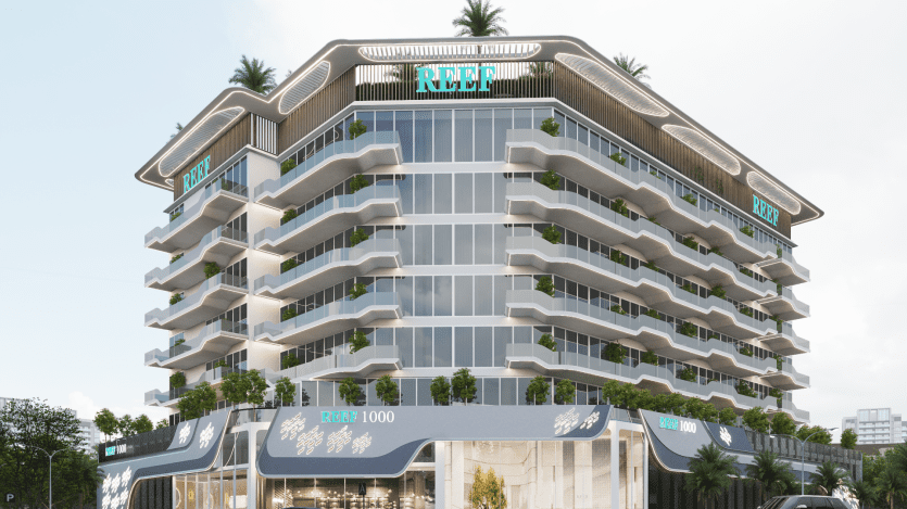 Un bâtiment moderne à plusieurs étages avec des balcons en verre et des plantes vertes à chaque étage. La structure est étiquetée « Reef » et dispose de verdure sur le toit. La base du bâtiment porte une signalisation indiquant « Reef 1000 ». Des arbres et un ciel dégagé sont visibles.