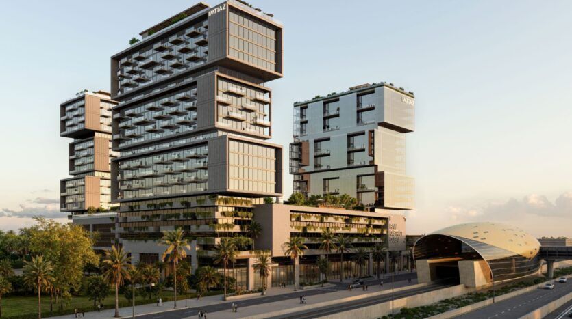 Des gratte-ciel modernes aux façades de verre empilées et en surplomb se dressent à côté d'une station de métro futuriste de forme ovale près de Raw District by Imtiaz ; des palmiers bordent la rue en face, avec quelques personnes marchant à proximité.