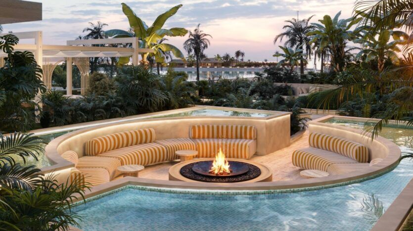 Des sièges incurvés, dotés de coussins rayés de jaune, entourent un foyer, aménagé dans un coin salon en contrebas au sein de la piscine carrelée du Château Piétrus, avec en arrière-plan une végétation tropicale luxuriante et des palmiers, sous un ciel aux teintes pastel au coucher du soleil.