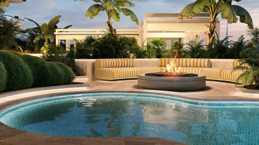 Une piscine aux lignes courbes se trouve face à un foyer circulaire, entourée de sièges rembourrés à rayures jaunes et de plantes tropicales luxuriantes, notamment des palmiers, créant ainsi, à la tombée de la nuit, un cadre extérieur à la fois relaxant et luxueux — une ambiance qui rappelle l'élégance du Château Piétrus.