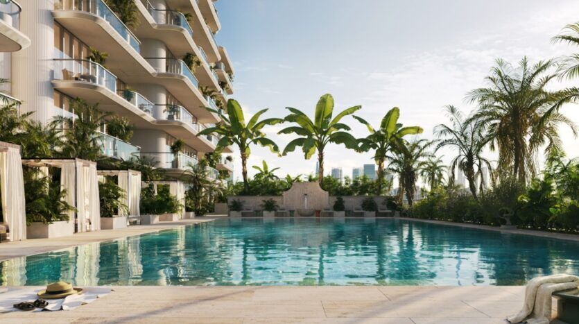 La luxueuse piscine du Château Piétrus est entourée de palmiers et de verdure ; on aperçoit, à gauche, des balcons modernes, des cabanes au bord de la piscine et, au-dessus, un ciel dégagé. Au premier plan, des serviettes, des chapeaux et un sac sont posés sur la terrasse de la piscine.
