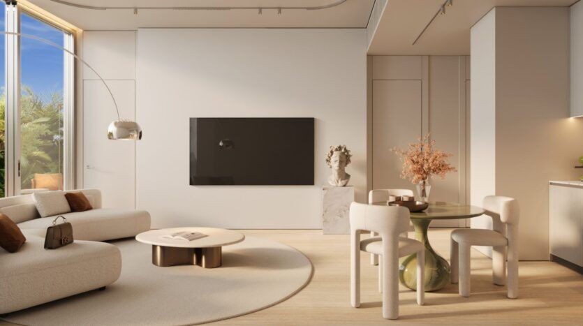 Ce salon minimaliste, aux tons neutres, avec sa table basse ronde, son canapé aux lignes courbes, sa table à manger en verre et ses chaises blanches, dégage une atmosphère de luxe discret qui rappelle Le Château Piétrus. L'espace comprend une télévision fixée au mur, un buste sculpté, une grande fenêtre laissant entrer la lumière naturelle et un vase orné de fleurs orange posé sur la table à manger.