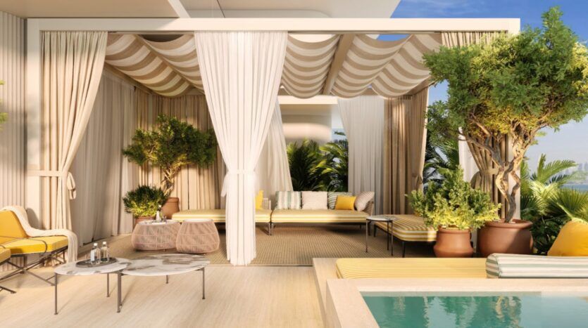 Une luxueuse cabane au bord de la piscine du Château Piétrus est agrémentée de coussins rayés jaunes et blancs, d'arbres en pot, de rideaux blancs et de tables en marbre, le tout sous un ciel ensoleillé.